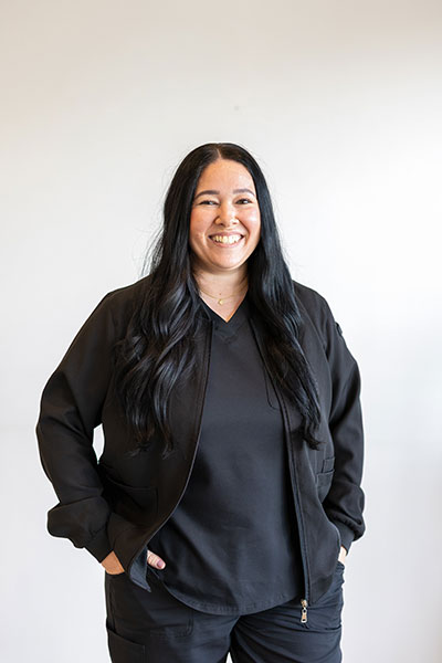 A woman with long dark hair stands confidently against a white background, wearing a black shirt and dark pants, radiating positivity with her smile.
