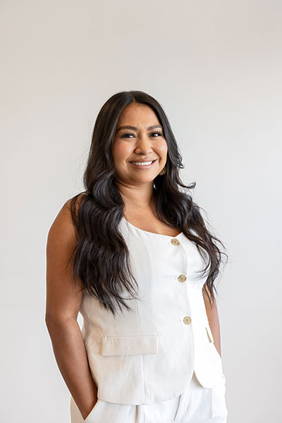 The image shows a woman with long hair standing against a white background. She has a broad smile and is wearing a light-colored top and dark pants.