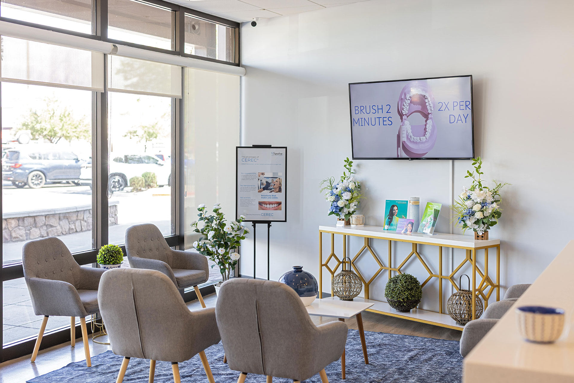 The image shows an interior space with a waiting area featuring modern furniture, including chairs and tables, arranged around a television displaying an advertisement. There are decorative elements such as potted plants, vases, and a framed sign on the wall. A large window allows natural light to enter the room, and there is a reception desk with a computer monitor in front of it. The setting suggests a professional or commercial environment, possibly a dental or medical office.