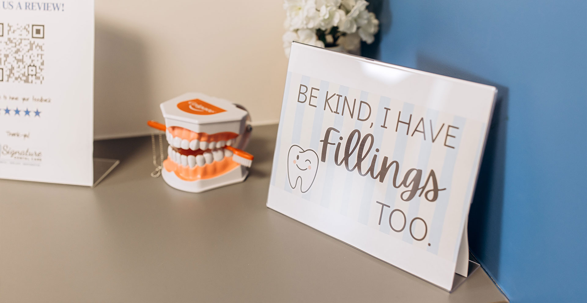 The image shows a whiteboard sign with handwritten text, placed on a desk next to a small gift item, against a blue wall.