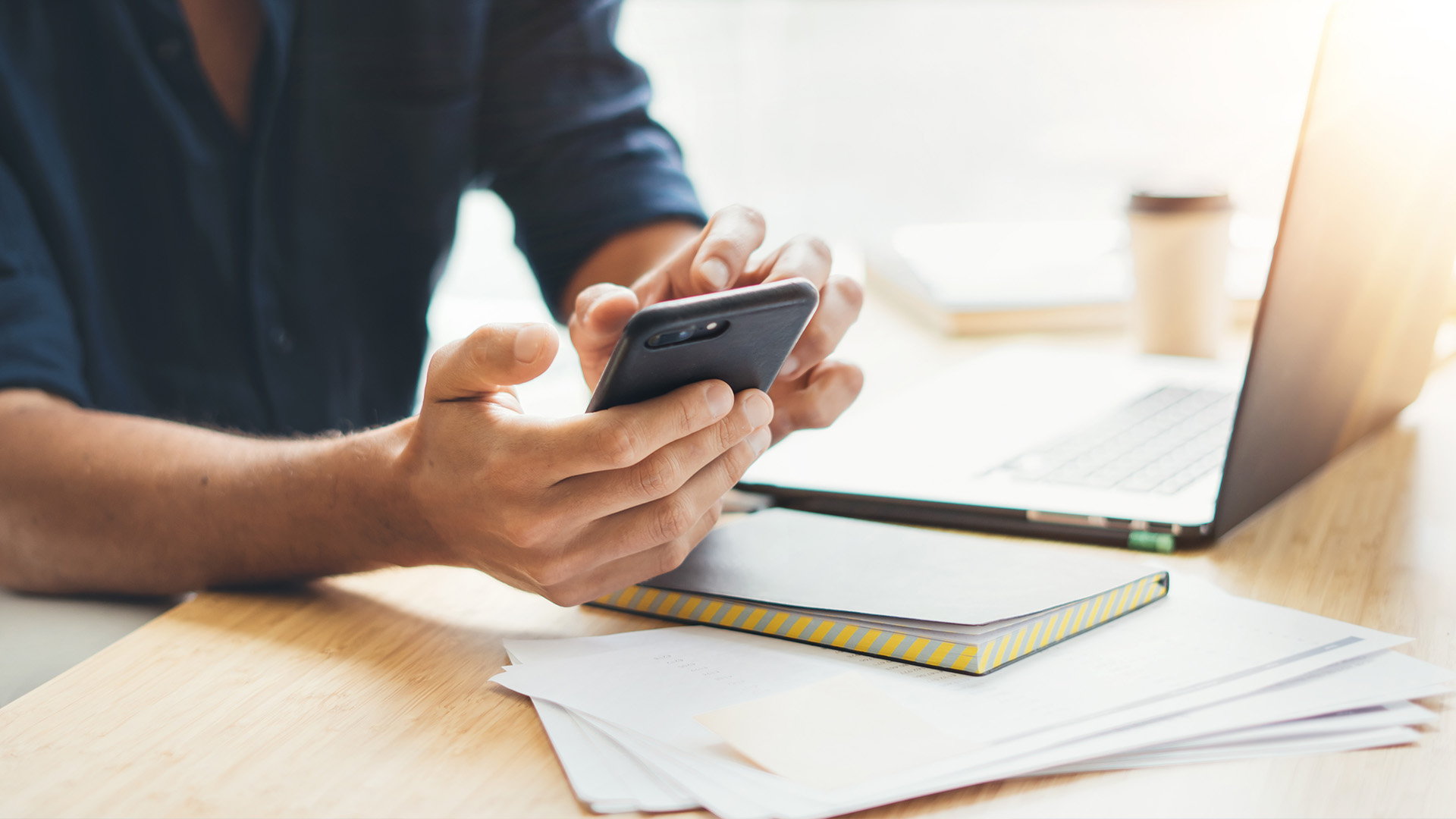 The image shows a person s hands holding a smartphone with a blurred background that includes a laptop, papers, and a desk in an office setting.