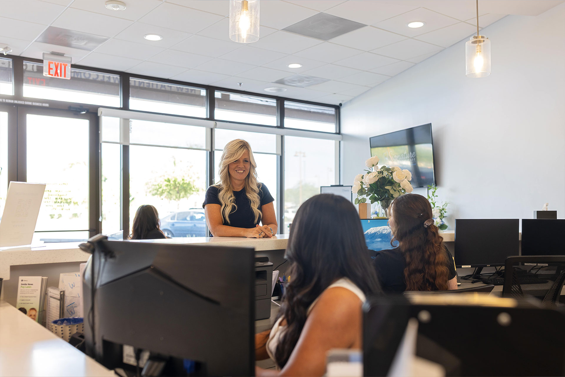 The image depicts an interior view of an office space with a woman standing behind a reception desk, engaged in conversation with two seated individuals.
