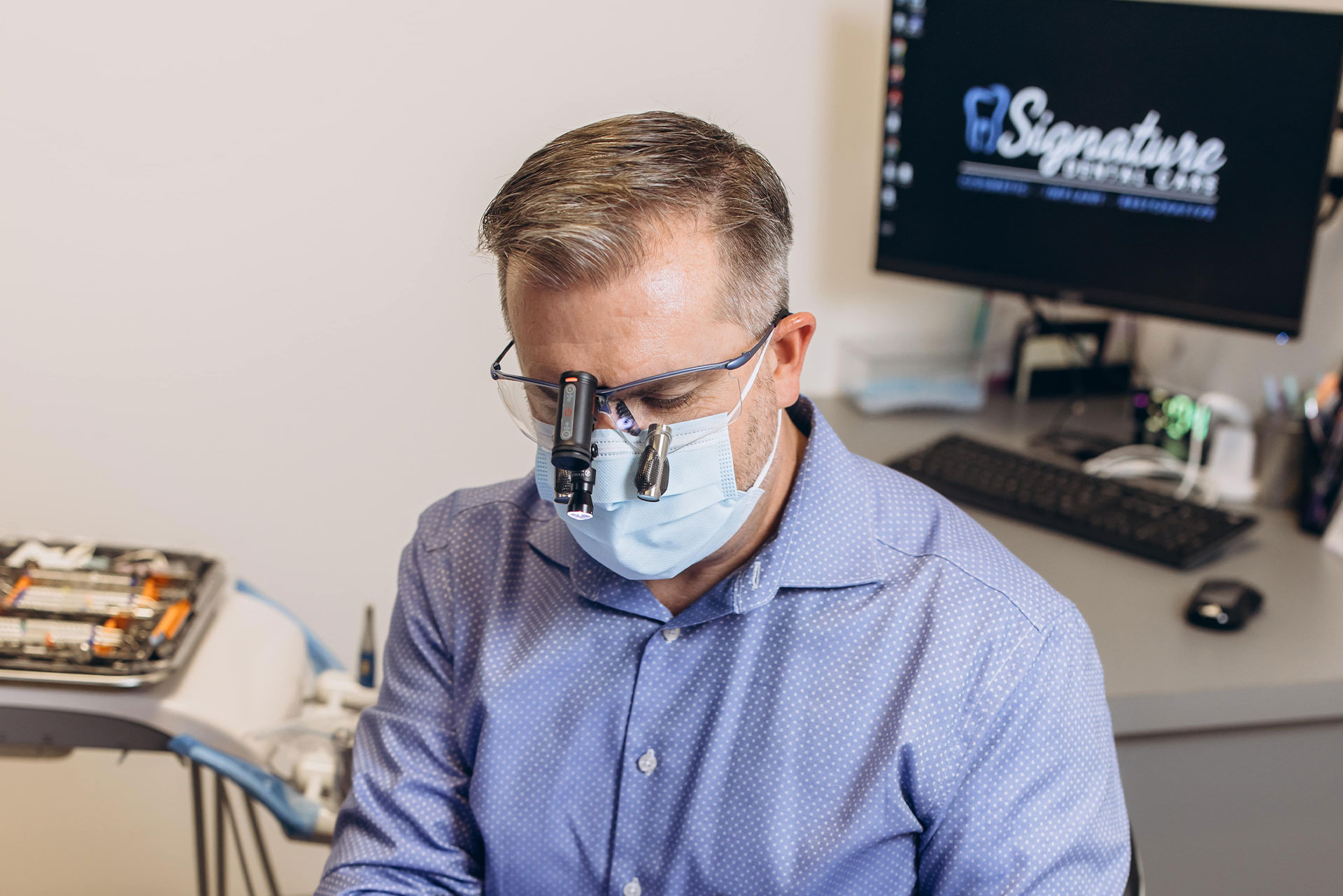 The image shows a man wearing a face mask and glasses, sitting at a desk with a computer monitor, keyboard, and mouse, working on a laptop.