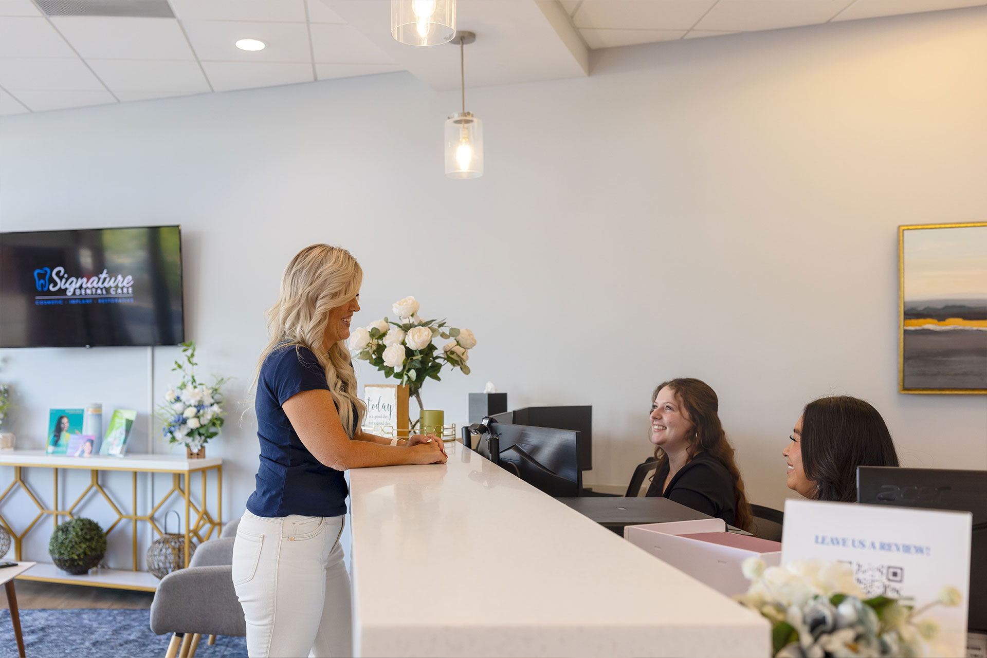 The image shows a woman standing behind a counter with a smiling expression, engaged in conversation with another person. They are both inside a well-lit room with modern decor, featuring a clean and minimalist aesthetic.