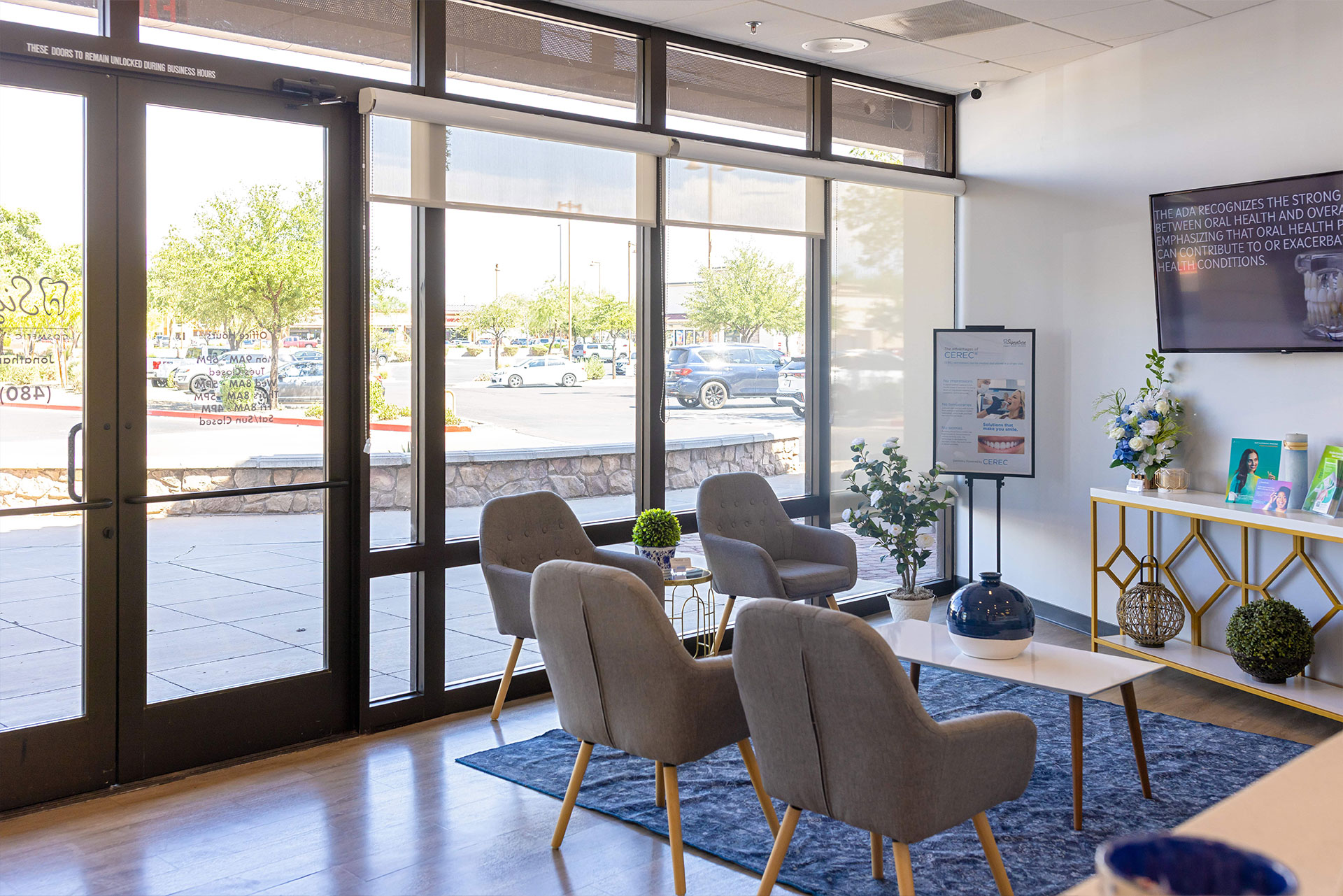 The image shows an interior space with large windows allowing natural light to enter, featuring modern furniture including chairs, a sofa, and tables, arranged around a central area with a television on the wall. There are potted plants and decorative items like vases and books, creating a welcoming atmosphere.