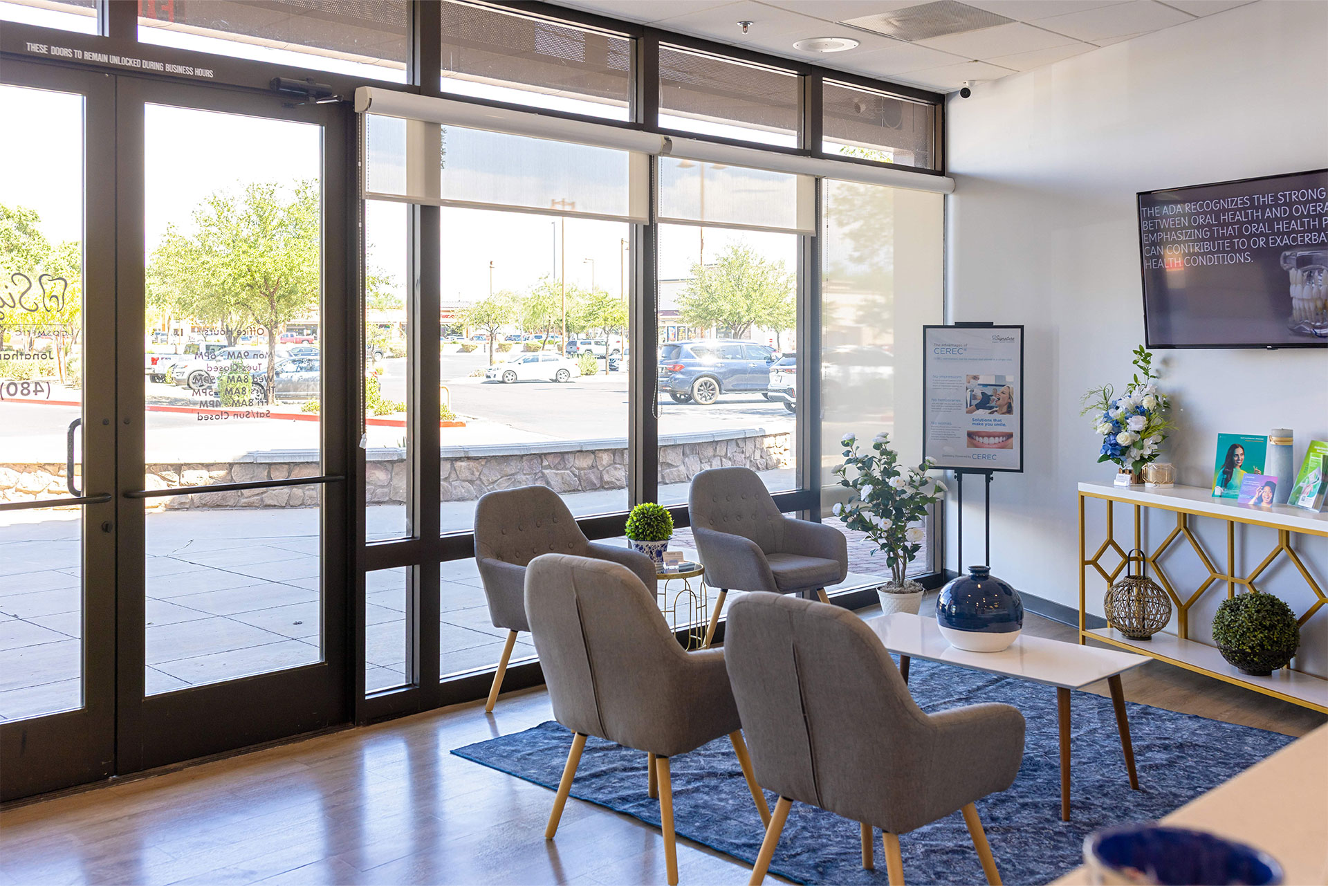 The image shows an interior space with large windows allowing natural light into the room. There is a modern furniture arrangement with a seating area featuring chairs and tables, a television mounted on the wall, and a reception desk with signage. A glass door leads to an outdoor patio area with additional seating and potted plants. The design suggests a contemporary and welcoming environment, possibly for a business or public space.
