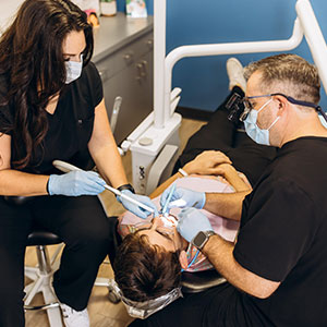 A group of dental professionals performing oral surgery on a patient, with the dentist using a drill and other instruments while seated at a chair.