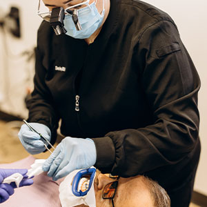 The image shows a person wearing a medical mask and gloves, performing dental work on a patient who is seated in a chair with their eyes closed.