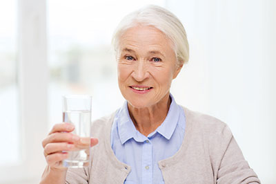 An elderly woman holding up a glass of water with her right hand, smiling and looking directly at the camera.