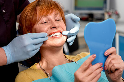 The image shows a woman sitting in a dental chair with a large model tooth being fitted onto her mouth by a dentist, who is holding up a blue dental impression tray.