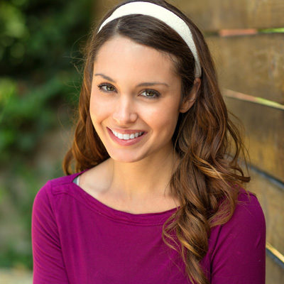 A woman with long brown hair wearing a purple top and a white headband smiles at the camera.