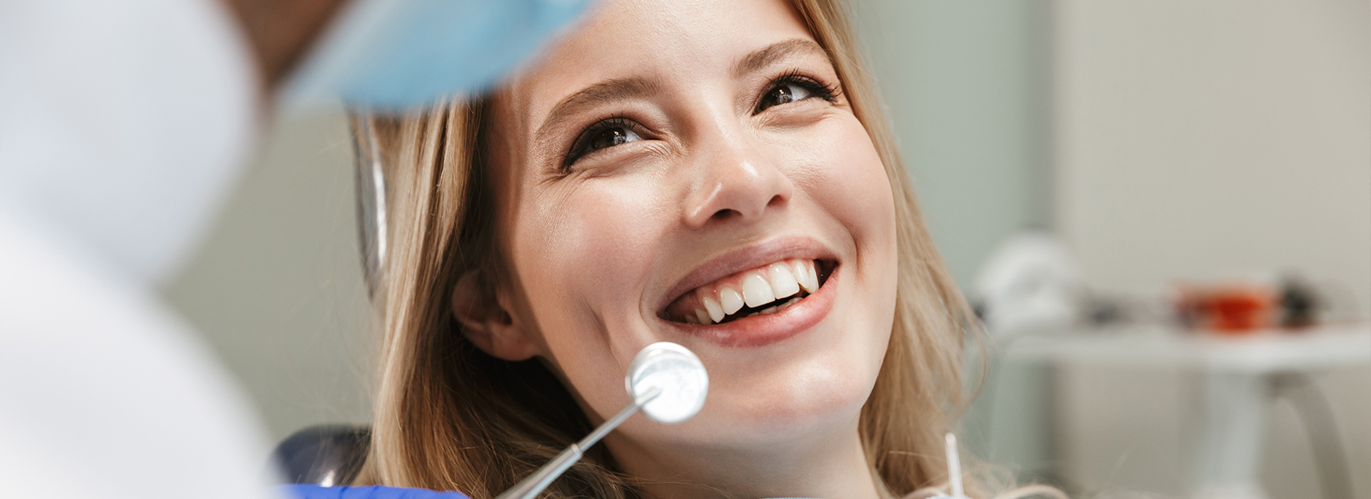A woman with a big smile is receiving dental care from a dentist wearing a white coat and stethoscope, with a medical setting visible in the background.