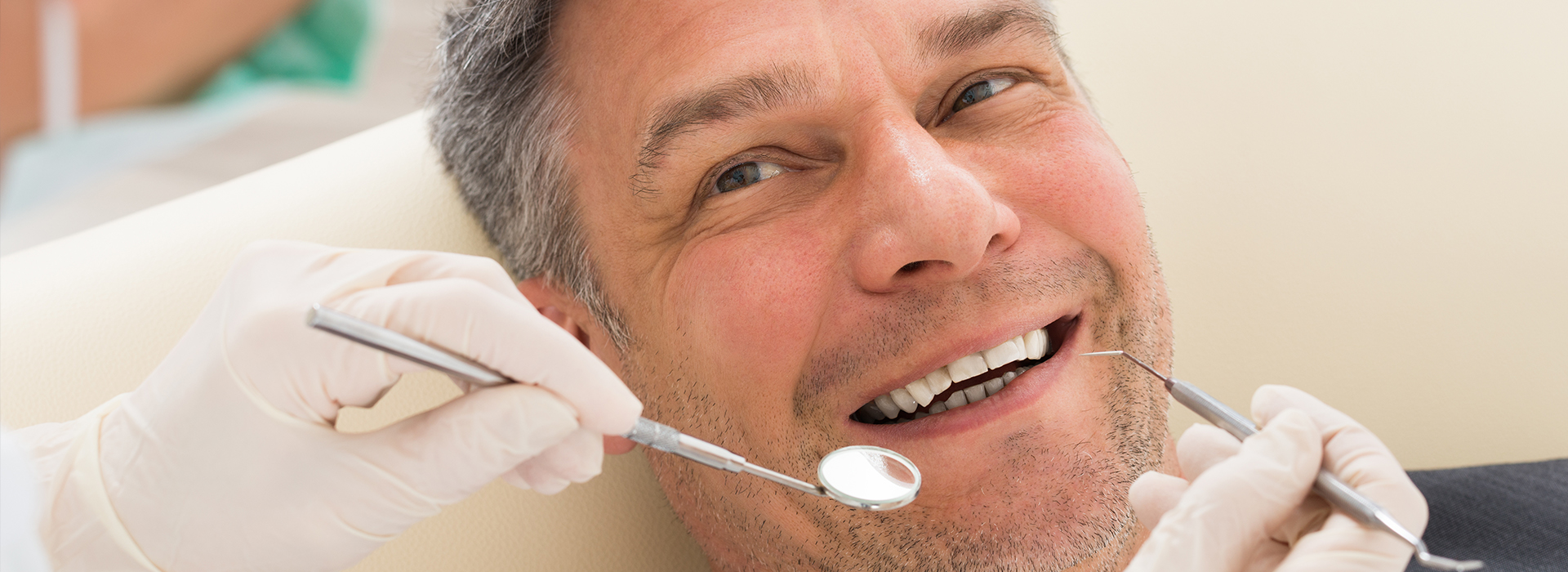 The image shows a man sitting in a dental chair with his mouth open, receiving dental treatment from a dentist who is working on his teeth using various tools visible around him.