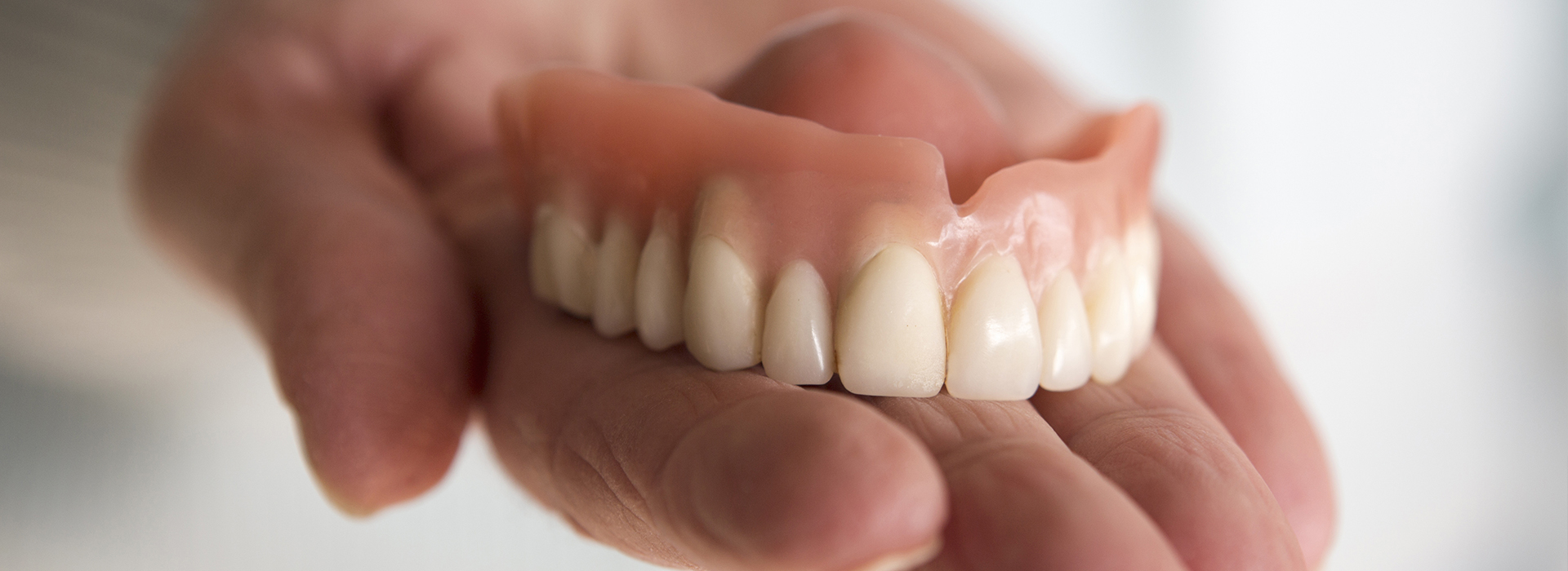 An elderly hand holding a set of dentures with yellowed teeth against a white background.