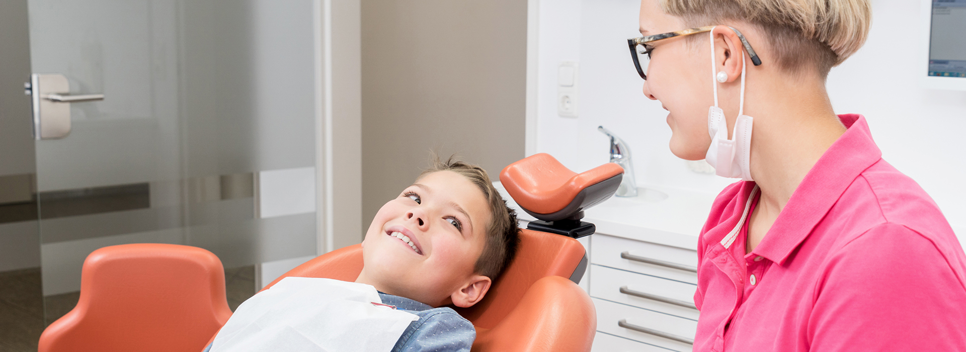 The image shows a dental office setting where a young boy is seated in a dental chair, smiling at the camera, with a woman standing behind him who appears to be a dental professional.