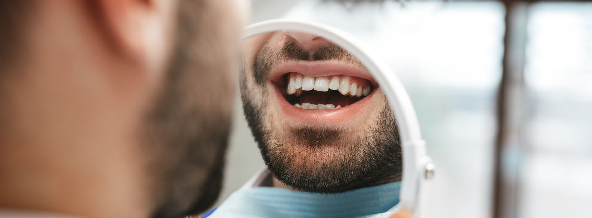 A man with a beard wearing a blue surgical mask and a white shirt has his mouth open, looking upwards, while sitting in front of a blurred background that appears to be an interior room with a window.