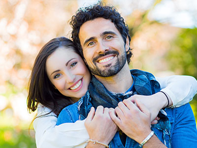 A man and woman are hugging each other, smiling and looking at the camera. They appear to be a couple enjoying a moment together outdoors during the day.