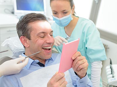 The image shows a man sitting in a dental chair with a smile, holding a pink cardboard sign that reads  Congratulations,  while a dentist and hygienist look on, suggesting a celebratory moment during a dental appointment.