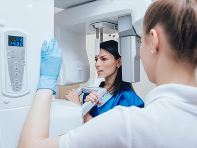 A woman stands next to a large 3D scanner machine, with another person behind her wearing a blue apron, inspecting the equipment.