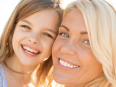 The image shows a woman with a child posing together for a photo, smiling and looking directly at the camera, set against a beach background under clear skies.