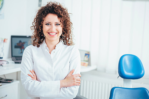 A smiling woman with curly hair stands confidently in an office setting, wearing a white blouse and standing beside a dental chair.