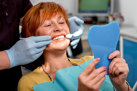 A woman sitting in a dental chair with a blue mouthguard on her teeth, smiling at the camera while holding up a mirror to show her teeth.