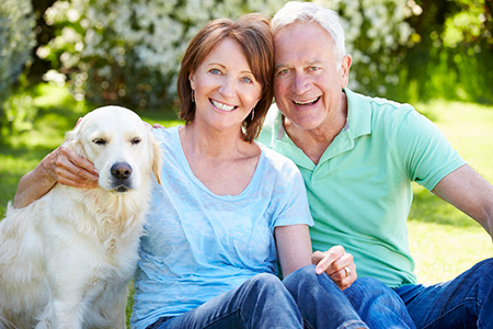 This image shows an older man and woman sitting outdoors on grass with a large yellow dog between them.