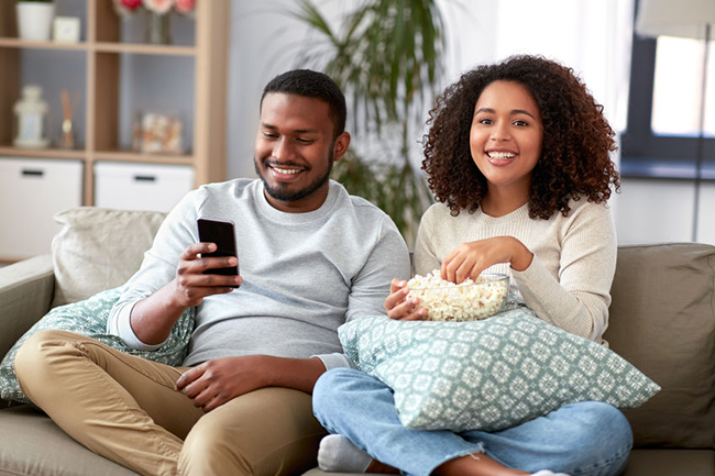This is a photograph of a man and a woman sitting on a couch, sharing a relaxed moment together while enjoying popcorn. The man is holding a smartphone, possibly browsing or texting. They appear to be in a comfortable domestic setting with modern interior design.