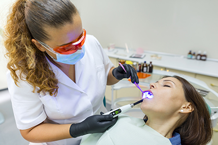 The image shows a dental hygienist performing oral care on a patient using a dental drill and an ultrasonic scaler, with both individuals wearing protective gear.