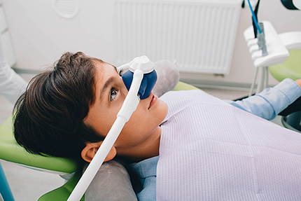 A young person wearing a medical mask, seated in a dental chair with their head positioned under a dental device, likely for a dental procedure.