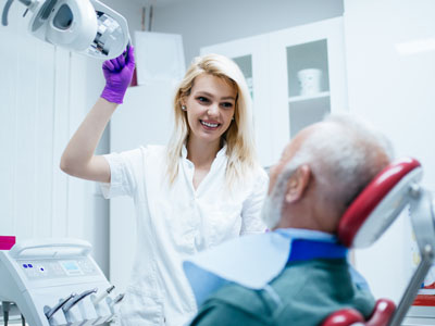 A woman in a white coat stands beside an older man with a medical mask on his face, both in a dental office setting.