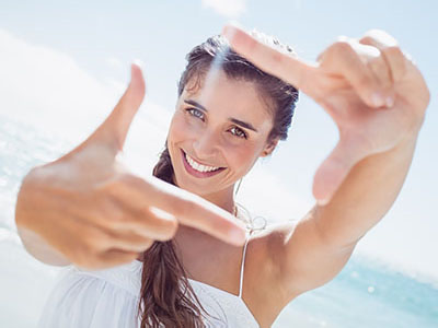 The image depicts a young woman taking a selfie with her hand held up to the camera lens, against a bright background that suggests a beach setting.